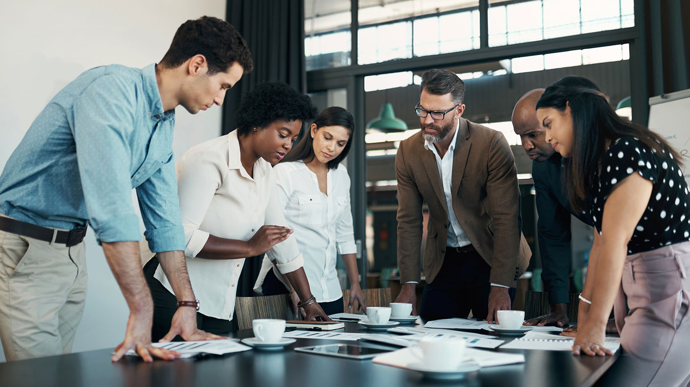 6 workers in an office stand over papers and documents on a table in a meeting room.