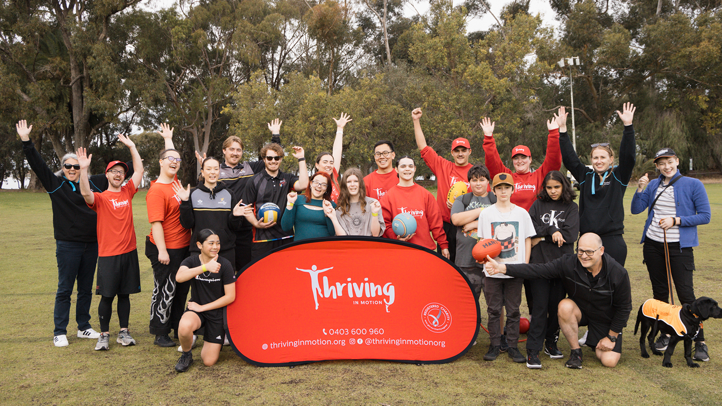 A group of people stand in a park with sports balls and banner that reads: Thriving in Motion.